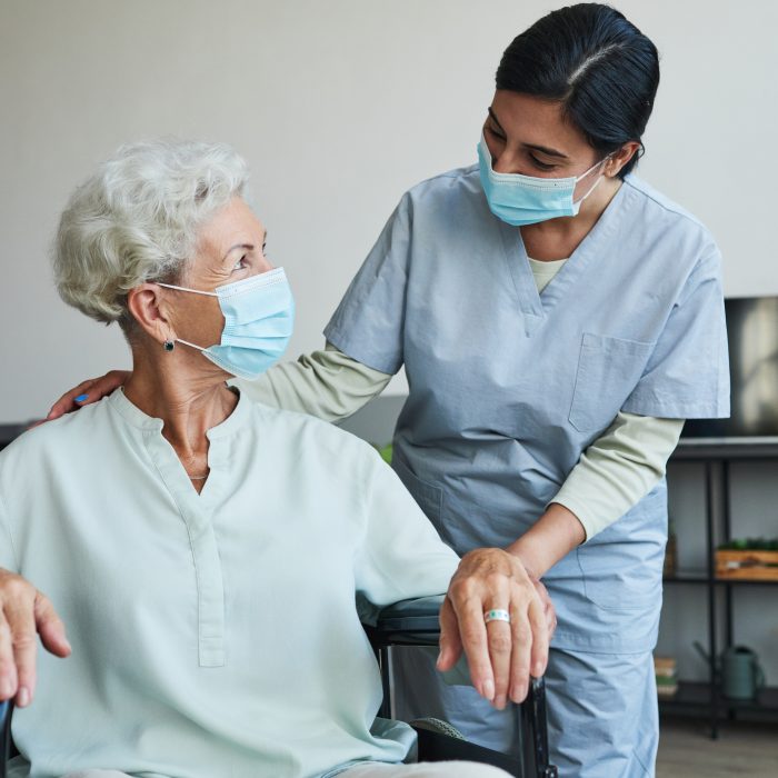 Portrait of female nurse assisting senior woman in wheelchair at home or in retirement center, both wearing masks